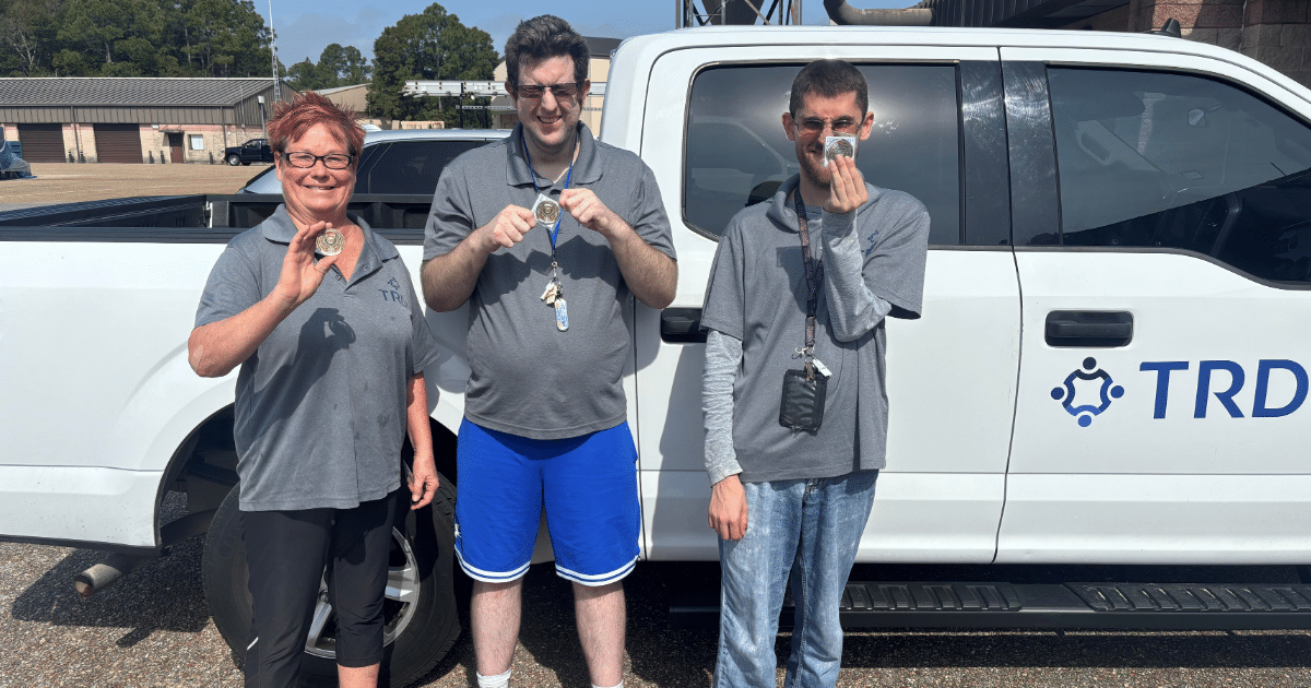 Three TRDI team members proudly displaying medals in front of a TRDI branded truck